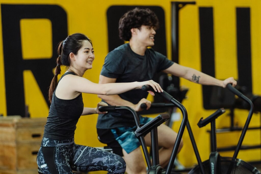 Asian man and woman enjoying a workout on stationary bikes in a gym, smiling and energetic.